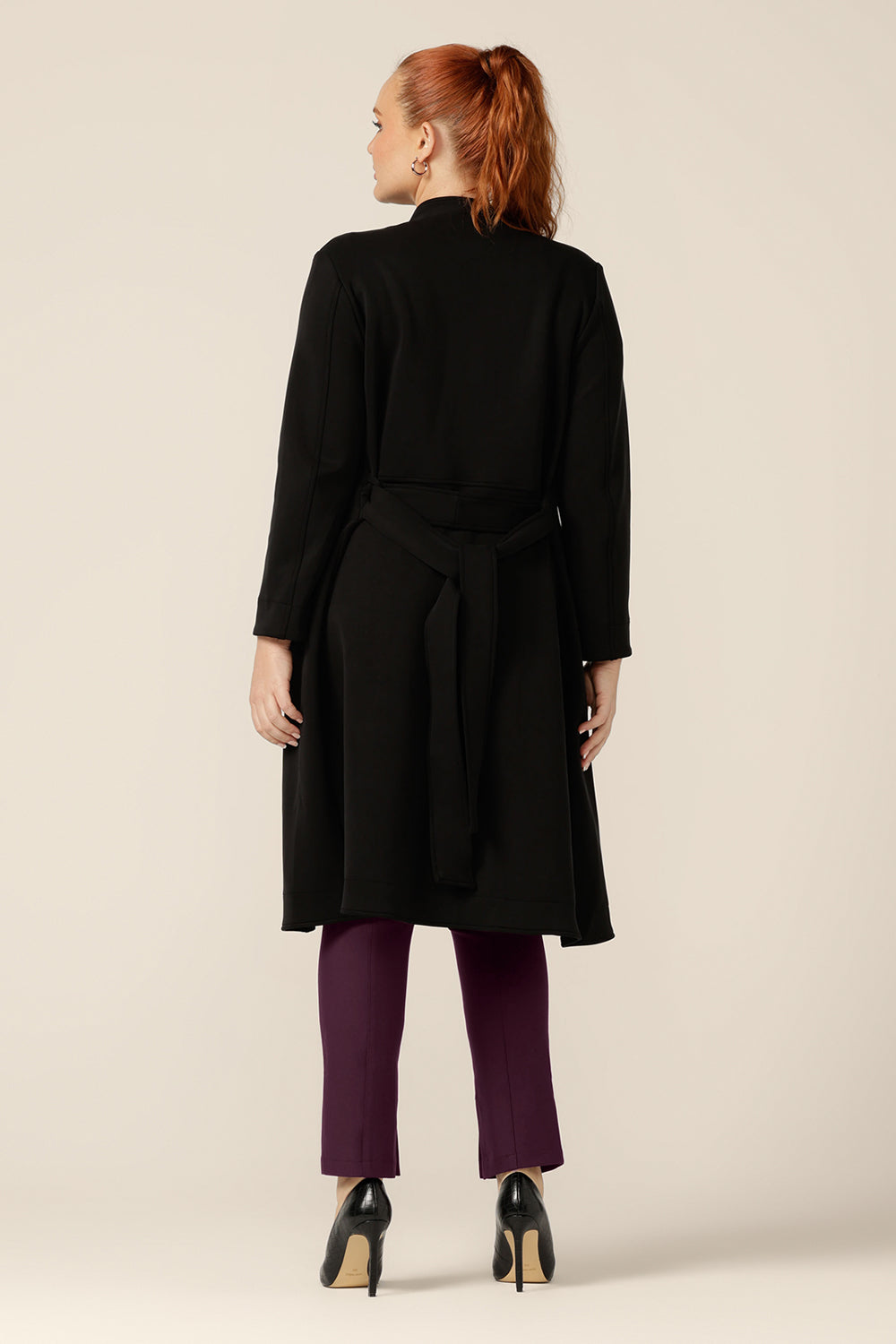 Back view of a curvy size, 12 woman wearing a softly tailored black trenchcoat in modal fabric. Worn with mulberry slim leg pants, this is a great coat for winter layering and travel. Shop in sizes 8 to 24.