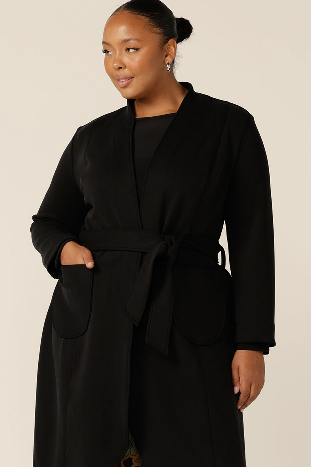 Great for chic city style, this black coat layers well for winter commuter workwear and as a travel coat. A curvy size 18 woman wears this softly tailored black trenchcoat in modal fabric tied at the waist.