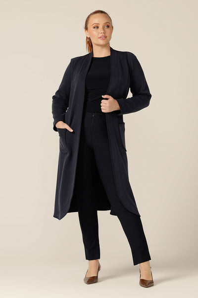 A size 12 woman wears the Marant Trenchcoat in Bluestone by Australian and New Zealand women's clothing company, L&F. A collarless, open fronted coat with tie belt and patch pockets, this classic navy coat is worn with a long sleeve, boat neck, navy top and slim-leg, navy trousers.