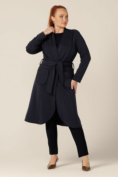 A size 12 woman wears the Marant Trenchcoat in Bluestone by Australian and New Zealand women's clothing company, L&F. Tied with a belt at the waist, this collarless coat features long sleeves and patch pockets. This classic navy coat is worn with a long sleeve, boat neck, navy top and slim-leg, navy trousers.