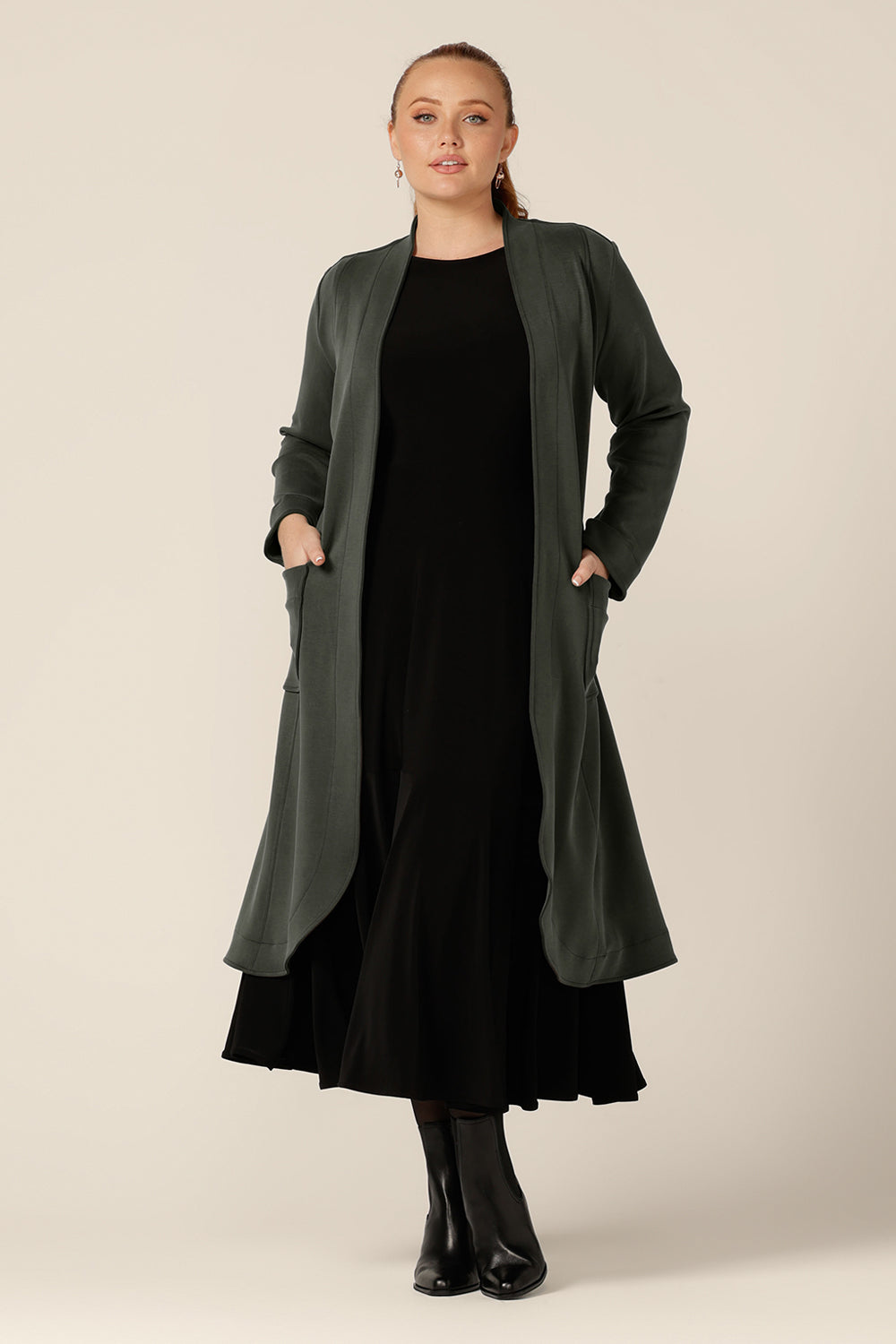 A size 12 woman wears the Marant Trenchcoat in Sage green by Australian and New Zealand women's clothing company, L&F. A collarless, open fronted coat with tie belt and patch pockets, this classic green coat is worn with a 3/4 sleeve, reversible black dress.
