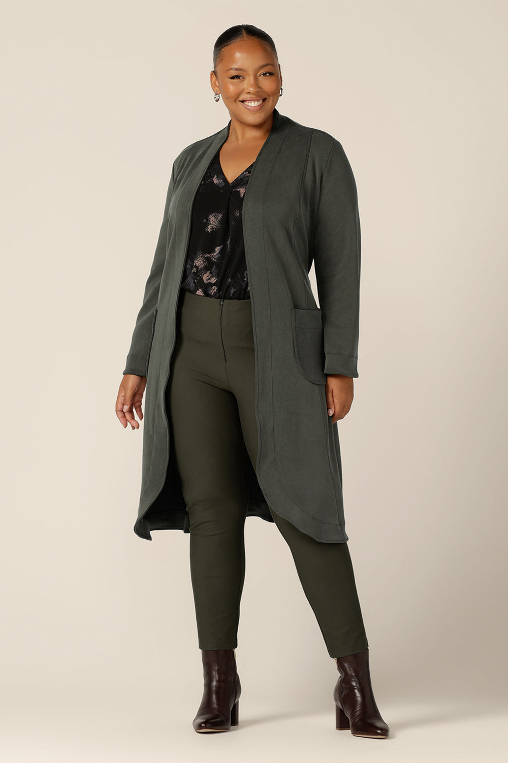 A classic winter coat for women, the Marant Trenchcoat in Sage green by Australian and New Zealand women's clothing label L&F is shown in a size 18, and worn with a printed, black jersey top and olive green, slim leg pants. A collarless, open fronted coat with tie belt and patch pockets, this women's winter jacket is available to buy in an inclusive 8 to 24 size range.