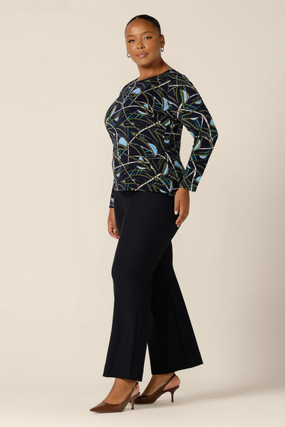 a size 18, plus size woman wears a long sleeve, boat neck top by Australian and New Zealand women's clothing brand, L&F. Featuring a blue, green and white print on a navy jersey base, this top is worn with navy, flared leg trousers for a complete workwear look. Available to shop in inclusive 8 to 24 sizes.