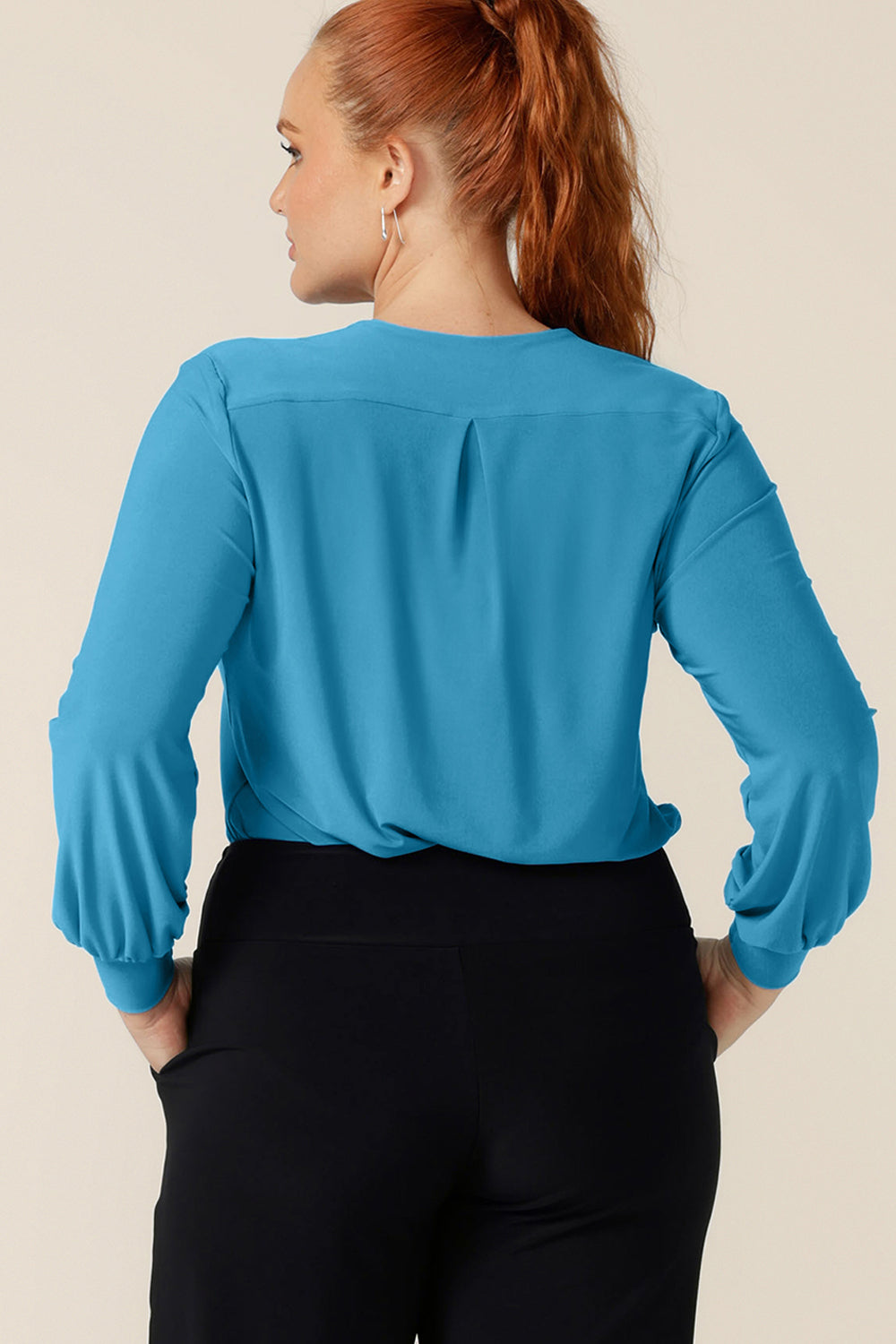 Back view of a curvy woman wearing a V-neck top with long bishop sleeves in Opal blue stretch jersey. Made in Australia by Australian and New Zealand women's clothing label, L&F, this workwear top is available to shop (with free shipping to New Zealand) in an inclusive size range of sizes 8 to 24.