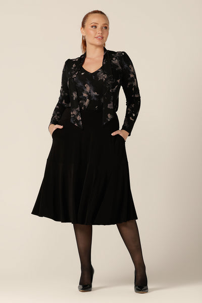 A curvy, size 12 woman wears a long sleeve, V-neck top with tie neck detail with a flared, knee-length black skirt. Both are by Australian and New Zealand women's clothing label, L&F.