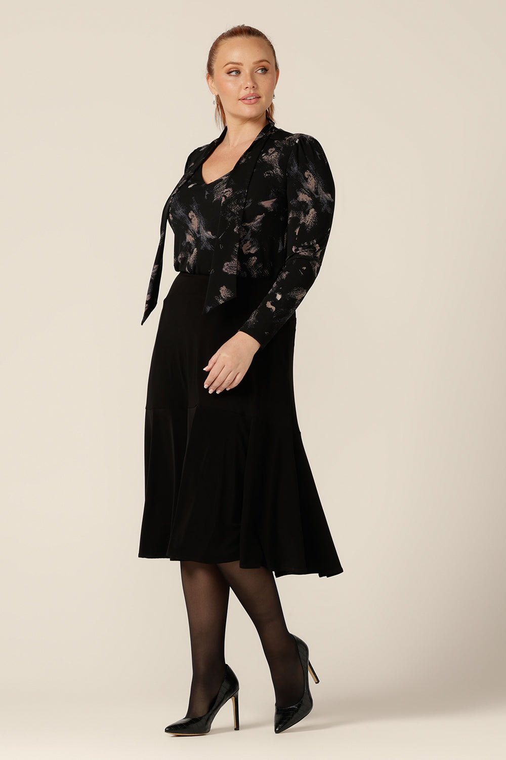 A curvy, size 12 woman wears a long sleeve, V-neck top with tie neck detail with a flared, knee-length black skirt. Both are by Australian and New Zealand women's fashion brand L&F and available to buy in an inclusive size range of 8 to 24.