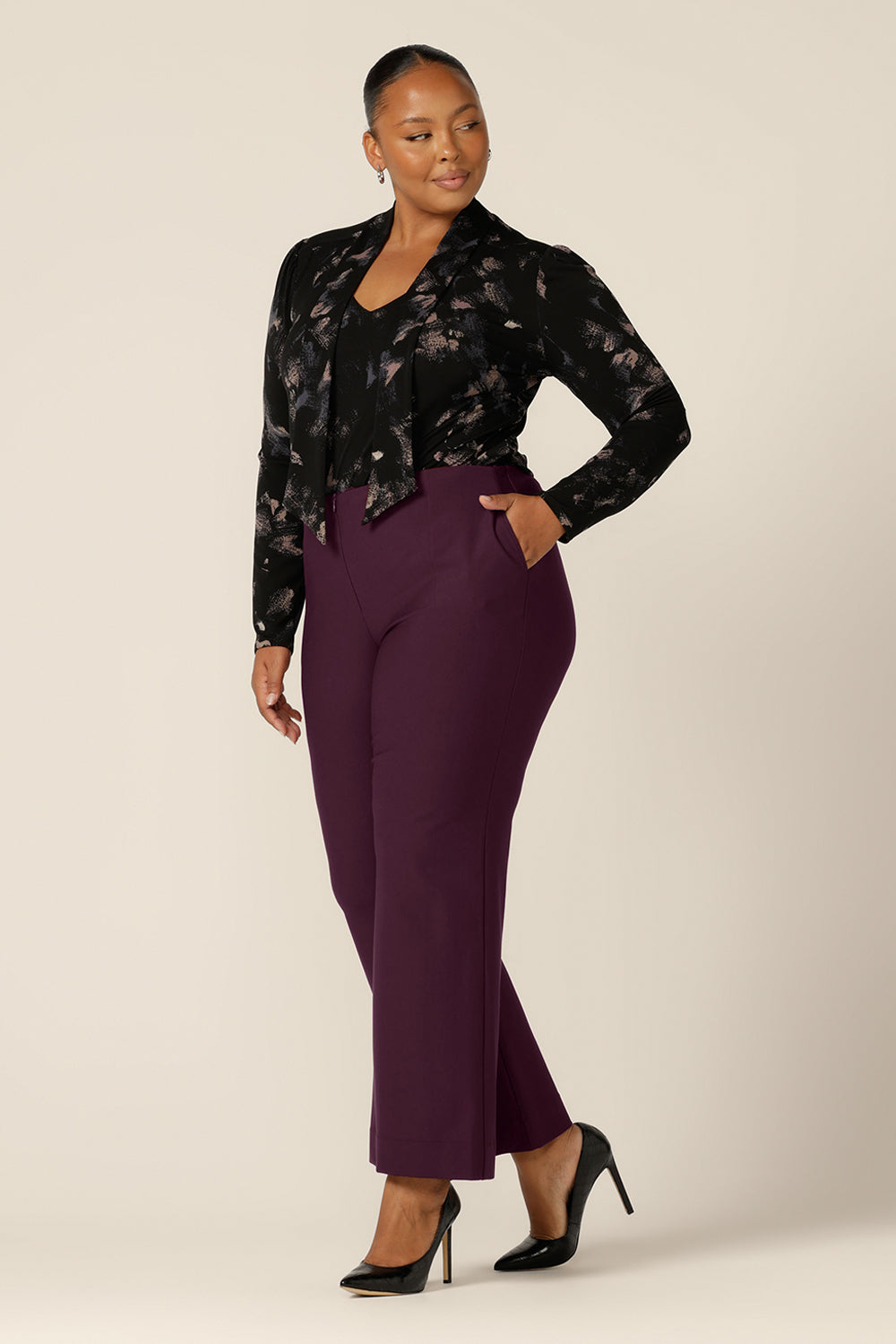 A fuller figure woman wears a long sleeve, V-neck top with tie neck detail in size 18 with flared leg, tailored pants in Mulberry. Both are by Australian and New Zealand women's clothing brand, L&F and are available to shop in an inclusive size range of sizes 8 to 24.