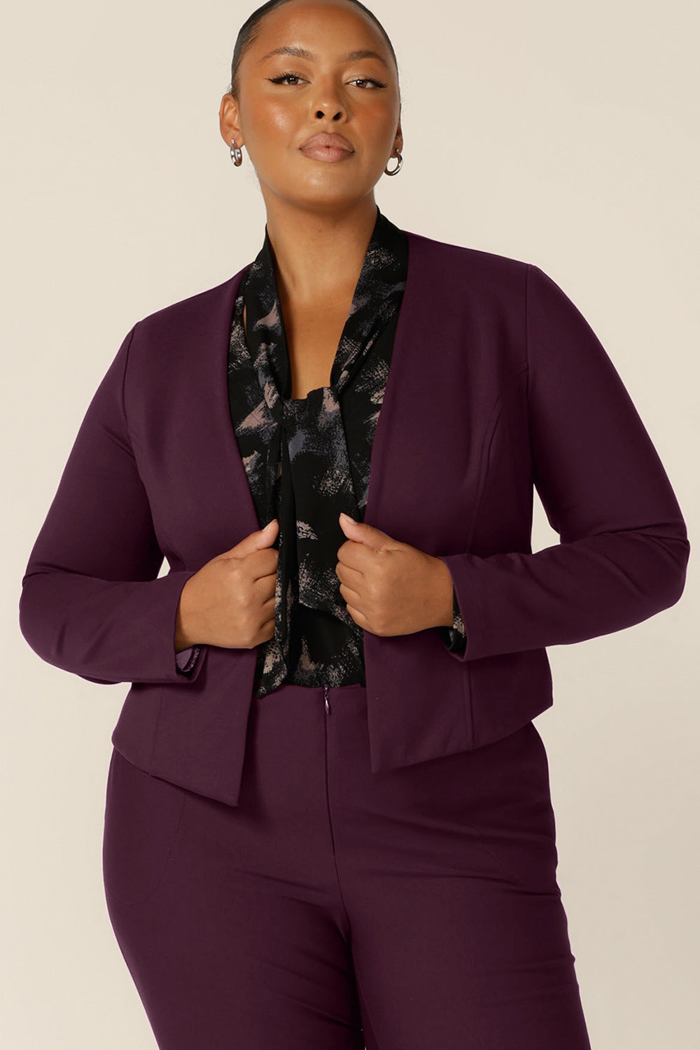 A plus size, size 18 woman wears a long sleeve, V-neck top with tie neck detail with flared leg, tailored pants in Mulberry and a collarless jacket in Mulberry for a strong work wear outfit.