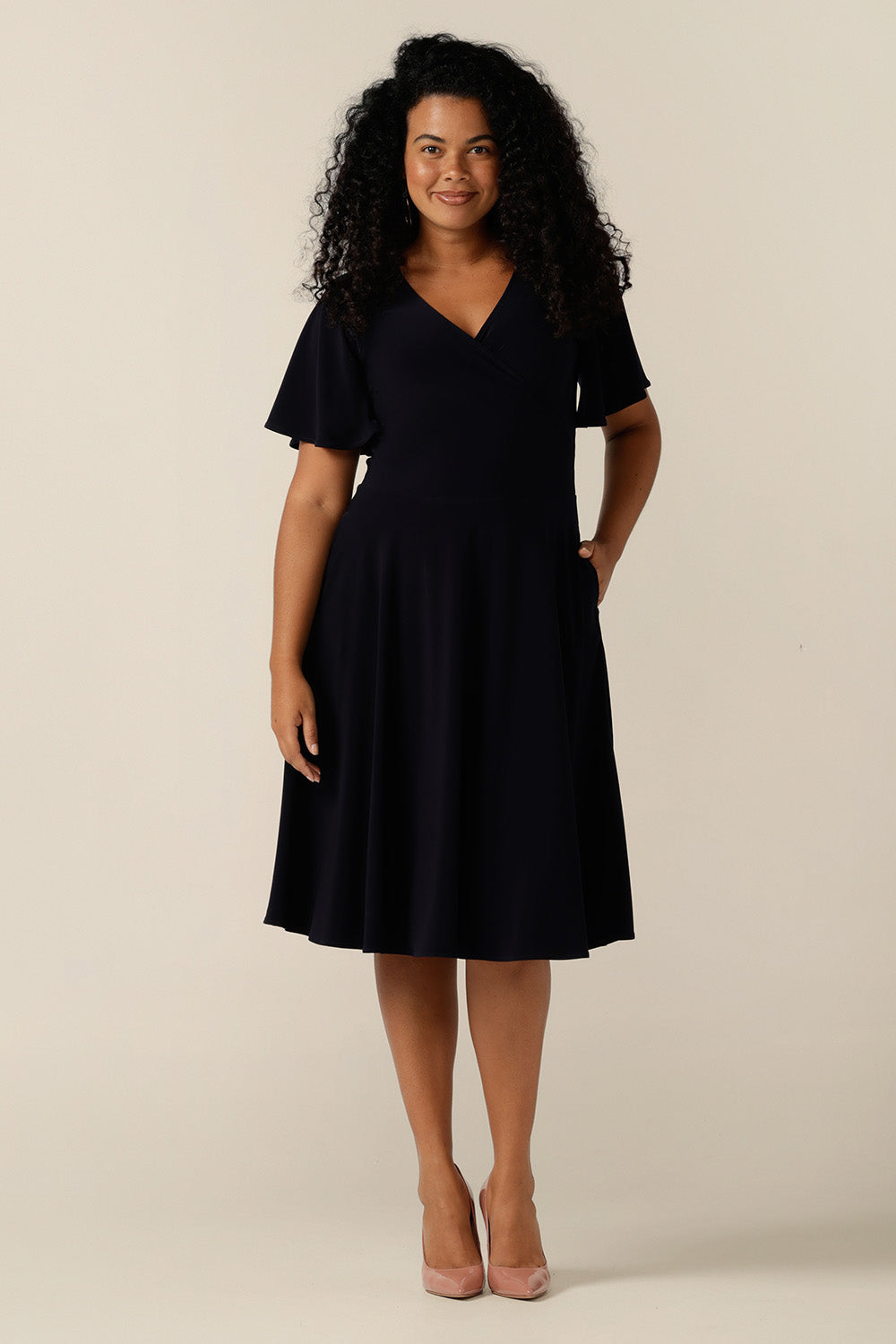 size 12, curvy woman wearing a reversible fixed wrap dress in navy jersey. The dress features flutter sleeves, knee length skirt and pockets. the reversible style of the dress allows her to be worn with a wrap front (as shown) or as a boat-neck dress. Designed as a work wear or evening dress, it is made in Australia for petite to plus sizes by women's clothing label, Leina and Fleur