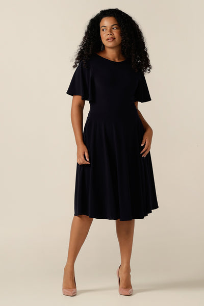size 12, curvy woman wearing a reversible fixed wrap dress in navy jersey. The dress features flutter sleeves, knee length skirt and pockets. the reversible style of the dress allows her to be worn with a wrap front or as a boat-neck dress. Designed as a work wear or evening dress, it is made in Australia for petite to plus sizes by women's clothing label, Leina and Fleur
