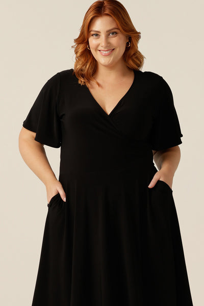 size 18, plus size woman wearing a reversible fixed wrap dress in black jersey. Worn with the wrap front forwards, she styles as elegant jersey little black dress. Featuring short flutter sleeves, a knee length skirt and pockets, this dress can be worn for work wear or going-out dress style. Made in Australia by women's clothing label, Leina and Fleur