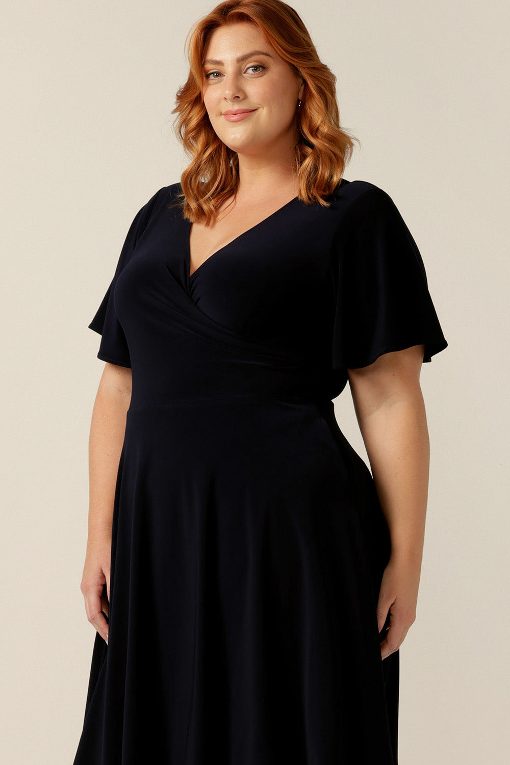 size 18, plus size woman wearing a reversible fixed wrap dress in navy jersey. The dress features flutter sleeves, knee length skirt and pockets. the reversible style of the dress allows her to be worn with a wrap front (as shown) or as a boat-neck dress. Designed as a work wear or evening dress, it is made in Australia for petite to plus sizes by women's clothing label, Leina and Fleur