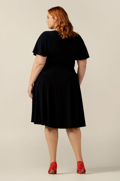 size 18, curvy woman wearing a reversible fixed wrap dress in navy jersey. The dress features flutter sleeves, knee length skirt and pockets. the reversible style of the dress allows her to be worn with a wrap front (as shown) or as a boat-neck dress. Designed as a work wear or evening dress, it is made in Australia for petite to plus sizes by women's clothing label, Leina and Fleur