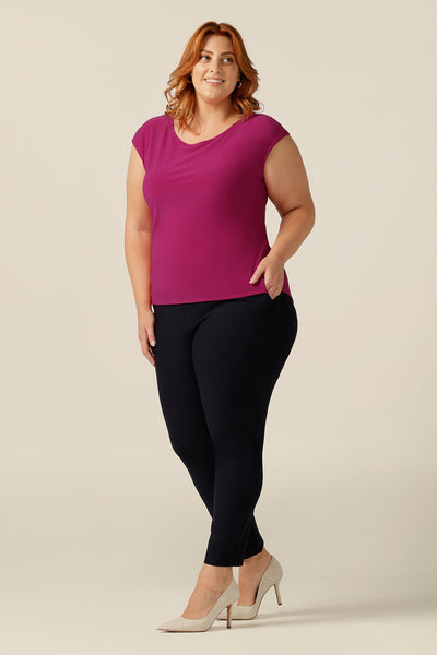 A  curvy, plus size woman wears a boat neck top with cap sleeves. The top is fuchsia pink stretch jersey, giving comfortable stretch to her slim fit top shape. Great as a work top, the pink Aspen Top is styled with slim-leg, tailored, work trousers in navy.