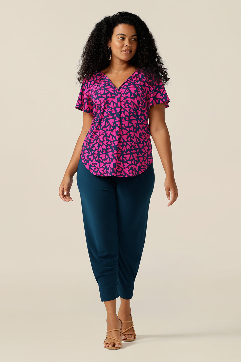 A size 12 curvy woman wears a V-neck short sleeve jersey top printed with pink hearts. She wears the top with cropped pants in blue jersey. Both are designed and made in Australia for petite to plus size women.