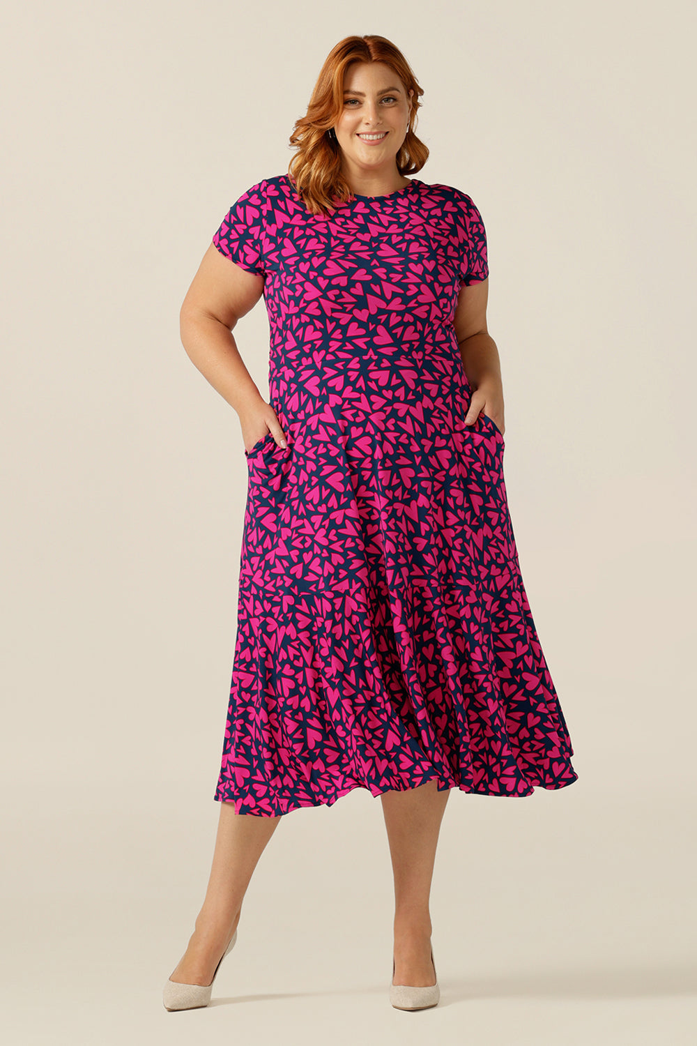 size 18 curvy woman wears a reversible jersey wrap dress. Worn with the crossover neckline to the front, this dress can be worn reversed with a boat neckline. This dress has short sleeves and a below-the-knee skirt with pockets. The dress is patterned with a pink heart print - perfect as a Valentines Day date-night dress or evening or occasionwear.