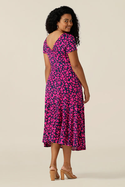 a size 12 curvy woman wears a reversible jersey wrap dress. Worn with the boat neckline to the front, this dress can be worn reversed with a crossover, V-neck. This dress has short sleeves and a below-the-knee skirt with pockets. The dress is patterned with a pink heart print - perfect as a Valentines Day date-night dress or evening or occasionwear.