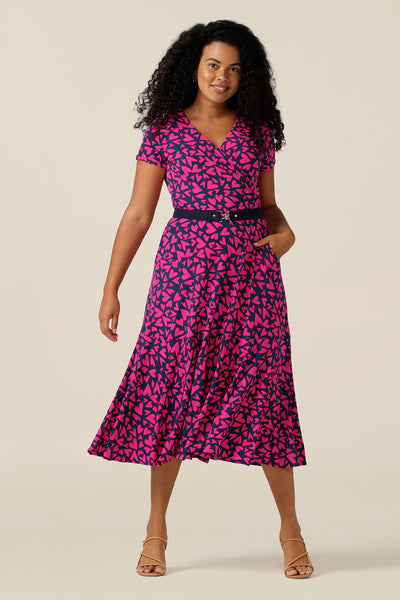 a size 12 curvy woman wears a reversible jersey wrap dress. Worn with the boat neckline to the front, this dress can be worn reversed with a crossover, V-neck. This dress has short sleeves and a below-the-knee skirt with pockets. The dress is patterned with a pink heart print - perfect as a Valentines Day date-night dress or evening or occasionwear.