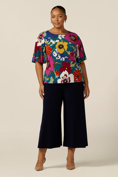 A fuller figure, size 18 woman wears a round neck jersey top with short flutter sleeves. She wears this Australian-made women's top over cropped-length, wide leg pants in navy for a corporate work wear look.