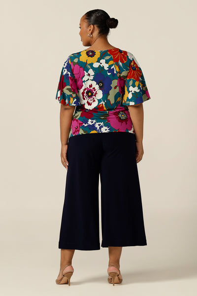 Back view of a size 18, plus size woman wearing a round neck , floral print, jersey top with wide flutter sleeves over wide leg, navy work pants . Australian-made, this quality work wear top gives feminine style to corporate pants and skirts. Shop this top online in sizes 8-24.