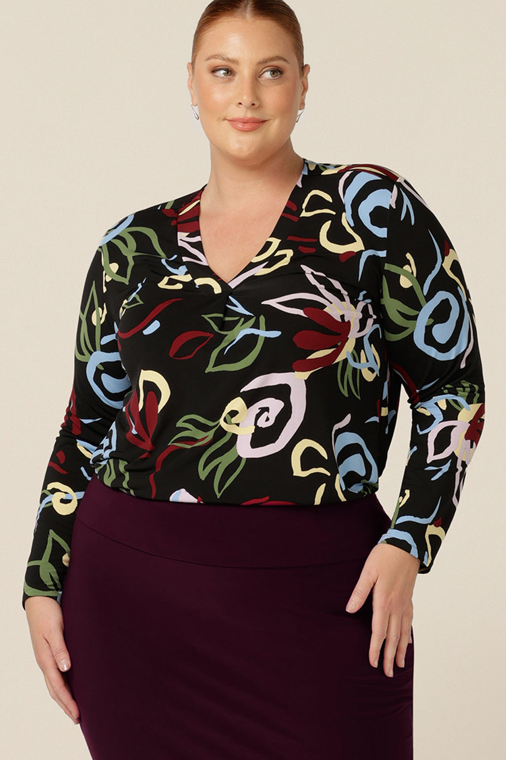 A fuller figure, size 18 woman wears a V-neck jersey top with long sleeves and a shirttail hemline. Australian-made by Australia and New Zealand women's clothing label, L&F, this long sleeve top features a black base abstract print.