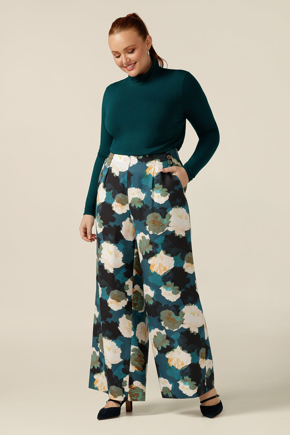 An Australian-made, women's long-sleeve, turtle neck top in petrol green bamboo jersey is worn by a curvy, size 12 woman with wide leg tailored pants for a workwear look. 