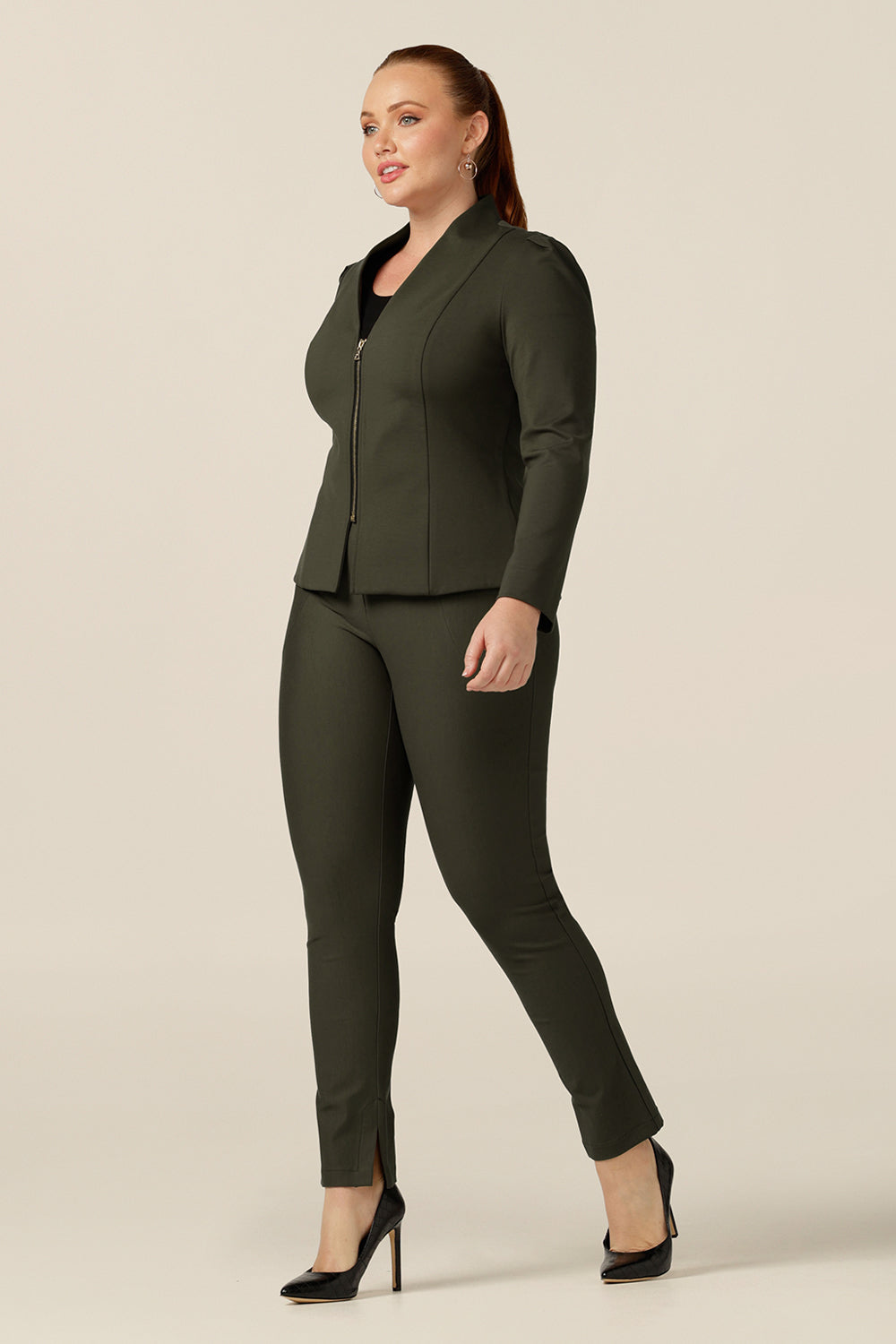 a size 12 curvy woman wears a tailored jacket with long sleeves, collarless V-neckline and zip fastening. Made in Australia in olive green ponte jersey, this is a quality-made, comfortable jacket is style with lim leg pants in olive green to create a soft suiting look for work and corporate wear.  