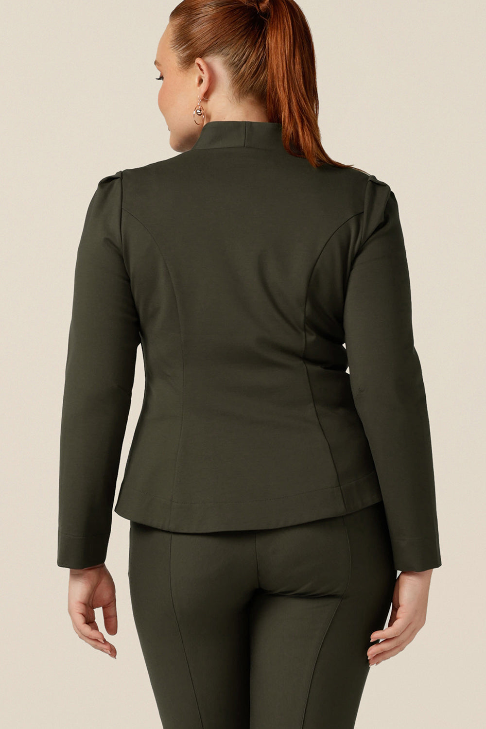 back view of a size 12 curvy woman wearing a tailored jacket with long sleeves, collarless V-neckline and zip fastening. Made in Australia in olive green ponte jersey, this is a quality-made, comfortable workwear jacket.