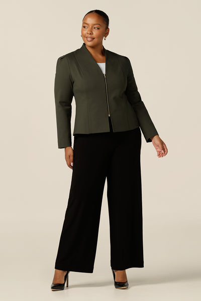 A size 18, fuller figure woman wears a tailored jacket with collarless V-neckline and zip fastening. In olive green ponte jersey, the workwear jacket is worn with wide-leg black pants for a corporate look. 
