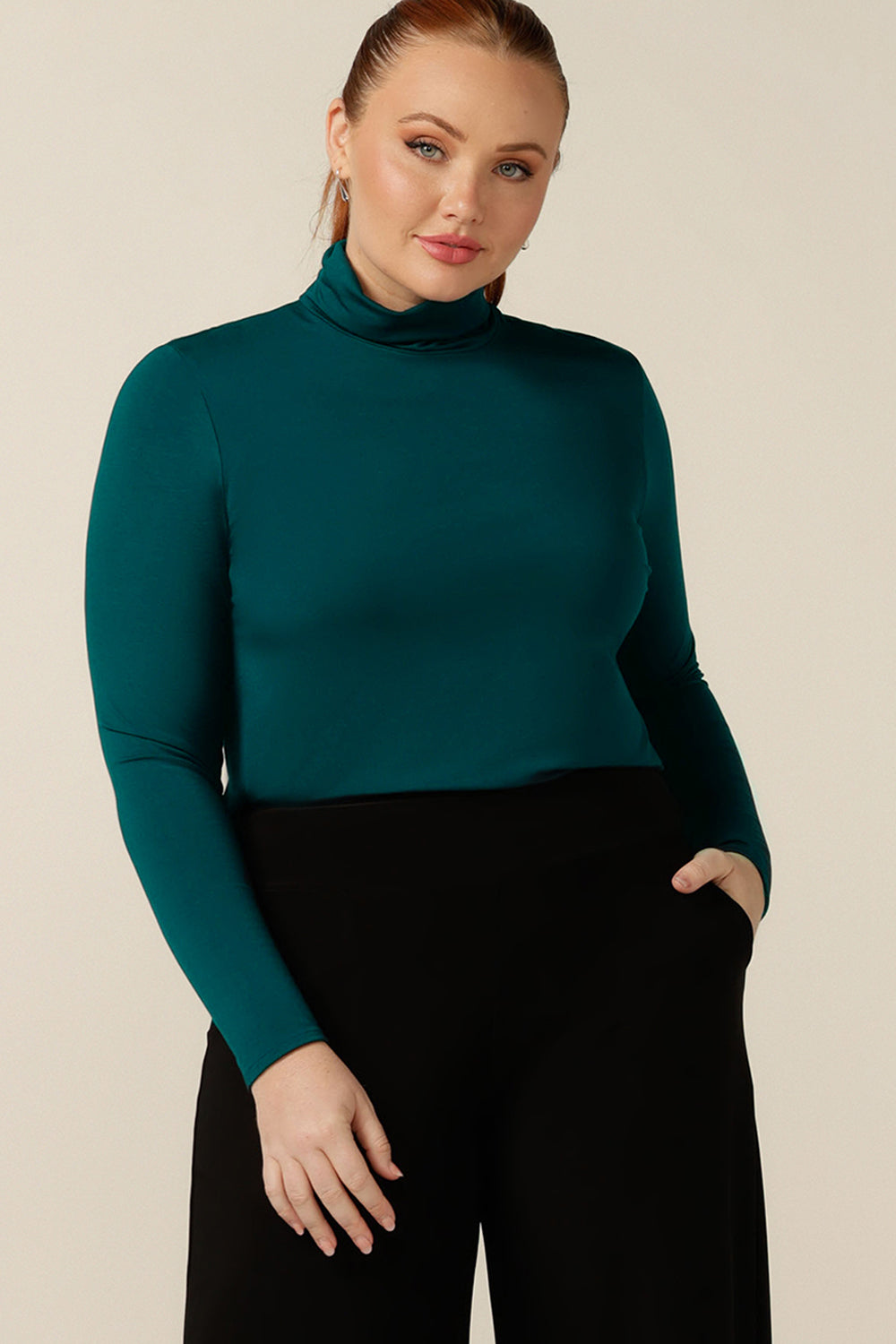 A size 12 curvy woman wears a long sleeve turtle neck top in Petrol green bamboo jersey. Made in Australia, this quality polo neck top styles for work and corporate wear.