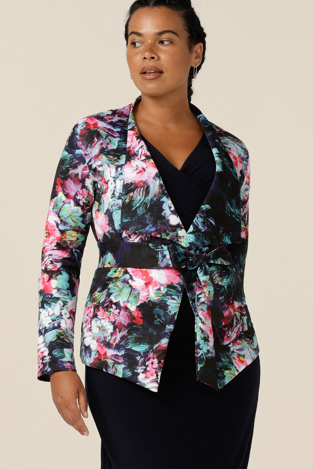 A size 12, curvy woman wears a wrap around jacket in comfortable stretch jersey. Patterned in an abstract floral print on a black base, this jacket has a V-neck, long sleeves and a belt tie. Designed as a work wear jacket and made in Australia, for the women of Australia and New Zealand, shop quality corporate jackets in sizes 8 to size 24 at Leina and Fleur.