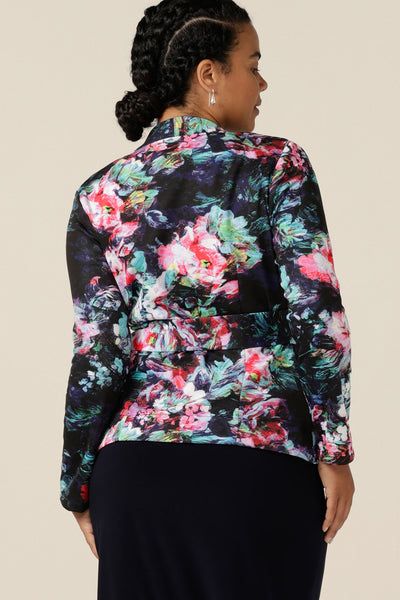 Back view of a size 12 woman wearing a wrap jacket in comfortable scuba jersey. Patterned in an abstract floral print on a black base, this jacket has a V-neck, long sleeves and a belt tie. Designed as a workwear jacket and made in Australia for the women of Australia and New Zealand, shop quality corporate jackets in sizes 8 to size 24 at Leina and Fleur.