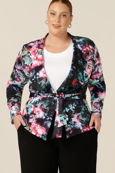 A size 18, plus size woman wears a comfortable work jacket in abstract floral print on a black base. This soft tailoring jacket has a tie belt, shown tied and long sleeves. Worn with black pants and a white T-shirt, this is a pretty jacket for work or weekend wear.
