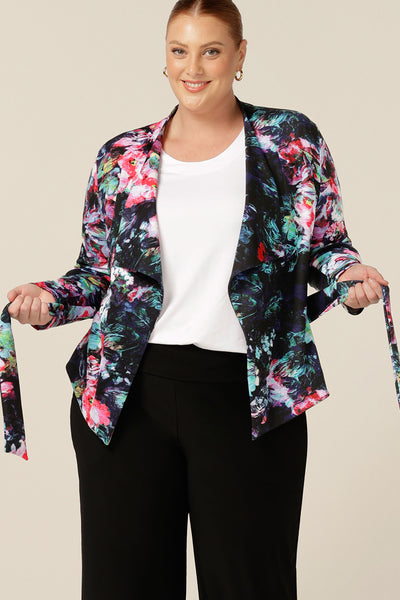 A size 18, fuller figure woman wears a comfortable work jacket in abstract floral print on a black base. This soft tailoring jacket has a tie belt, soft collar and long sleeves. Worn with black pants and a white T-shirt, this is a pretty jacket for work or weekend wear. Shop with free shipping to New Zealand..