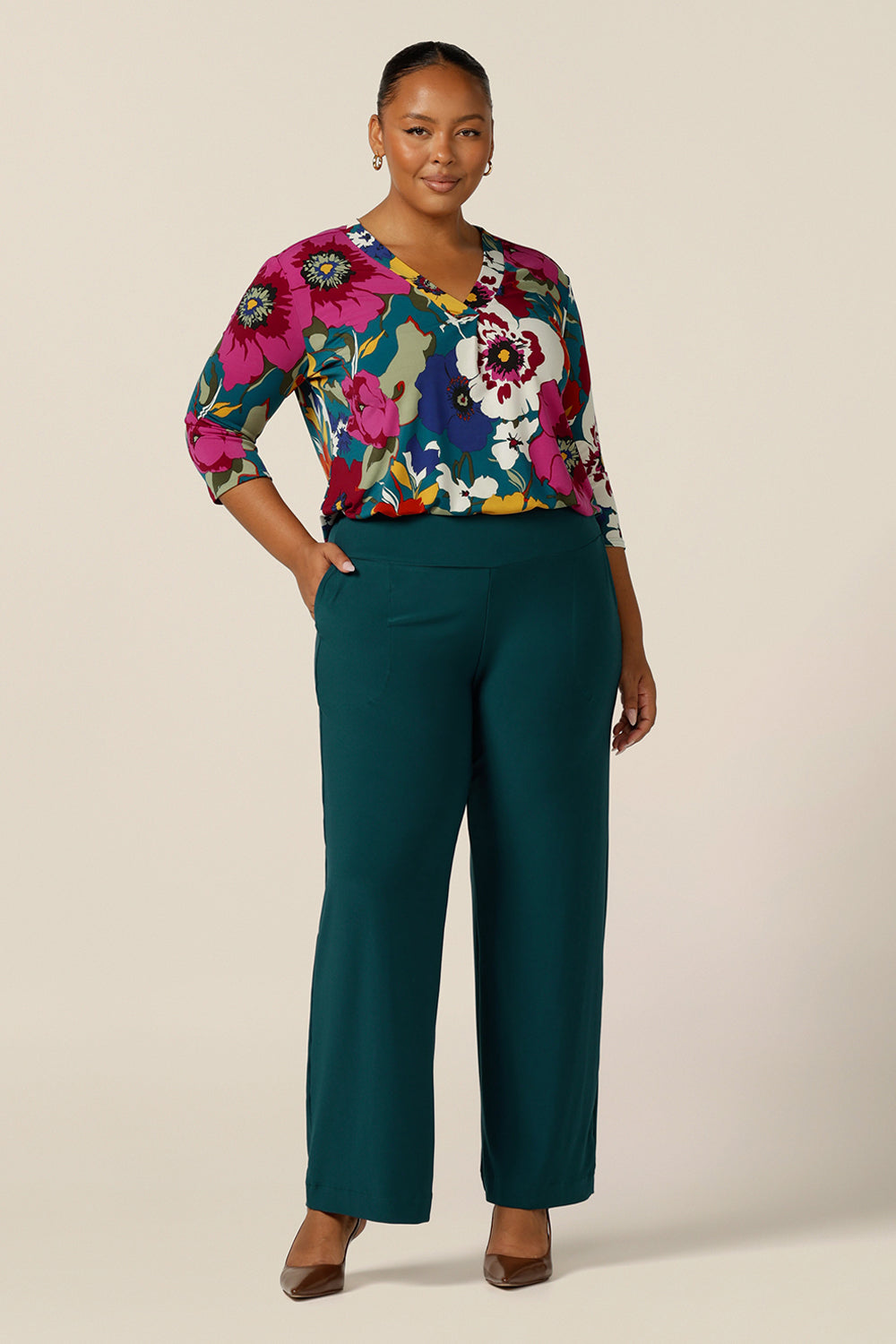 A size 18, plus size woman wears a jersey, V-neck top with 3/4 sleeves. A tailored top for work and corporate wear, the floral print top is tucked in to wide-leg trousers as a workwear outfit.