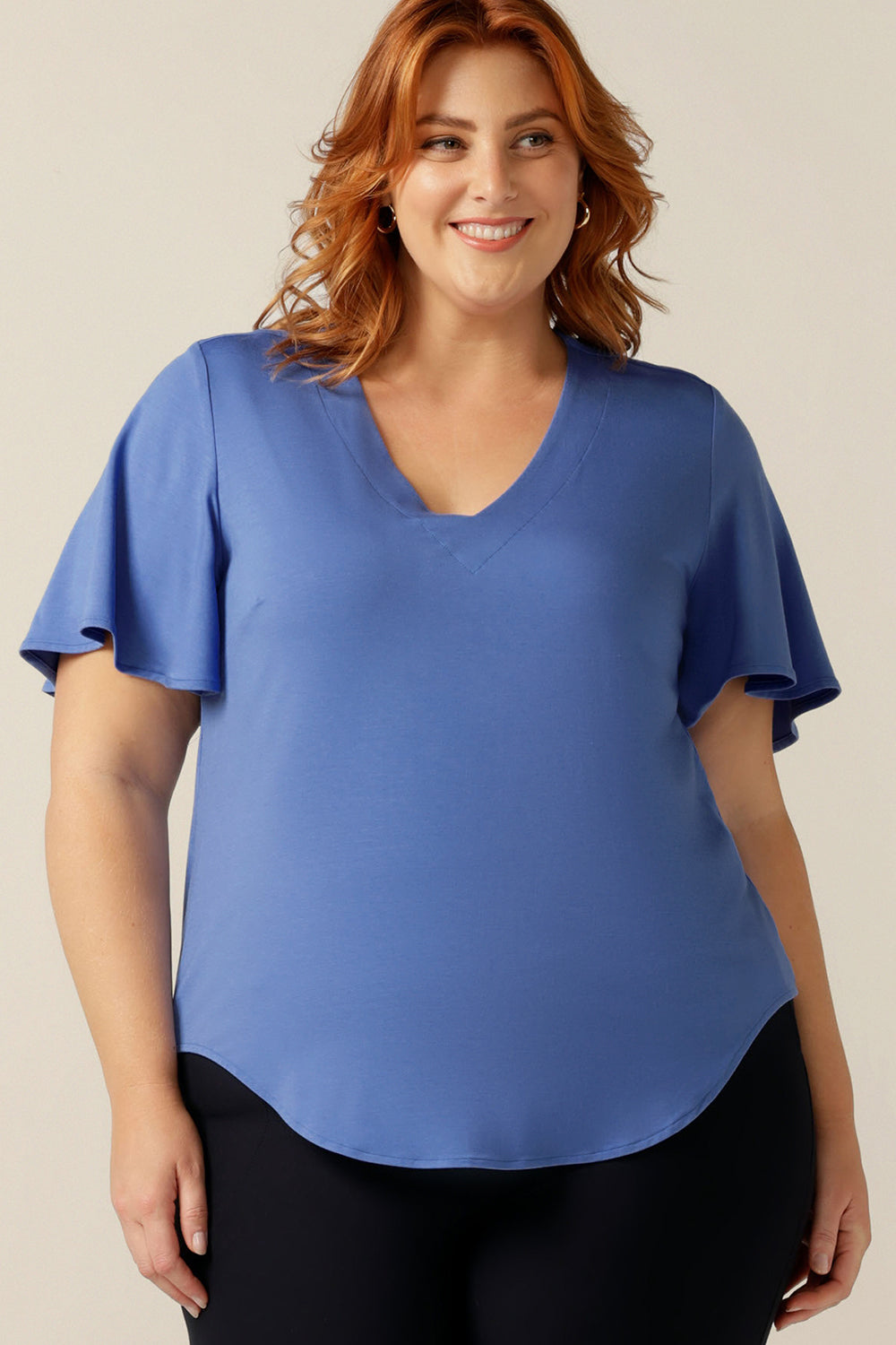 A plus size, size 18 woman wears a V-neck blue top with short flutter sleeves. Made from bamboo jersey, this is a natural fibre top that is lightweight, breathable and sustainable.  