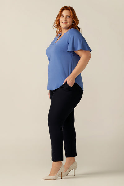 A plus size, size 18 woman wears a V-neck blue top with short flutter sleeves. Made from bamboo jersey, this is a natural fibre top that is lightweight, breathable and sustainable. The blue bamboo jersey top is worn over slim-leg black jersey trousers for a smart-casual look