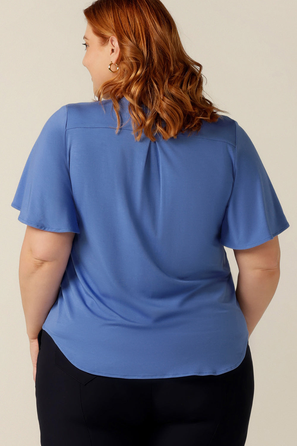 A plus size, size 18 woman wears a V-neck blue top with short flutter sleeves. Made from bamboo jersey, this is a natural fibre top that is lightweight, breathable and sustainable.  