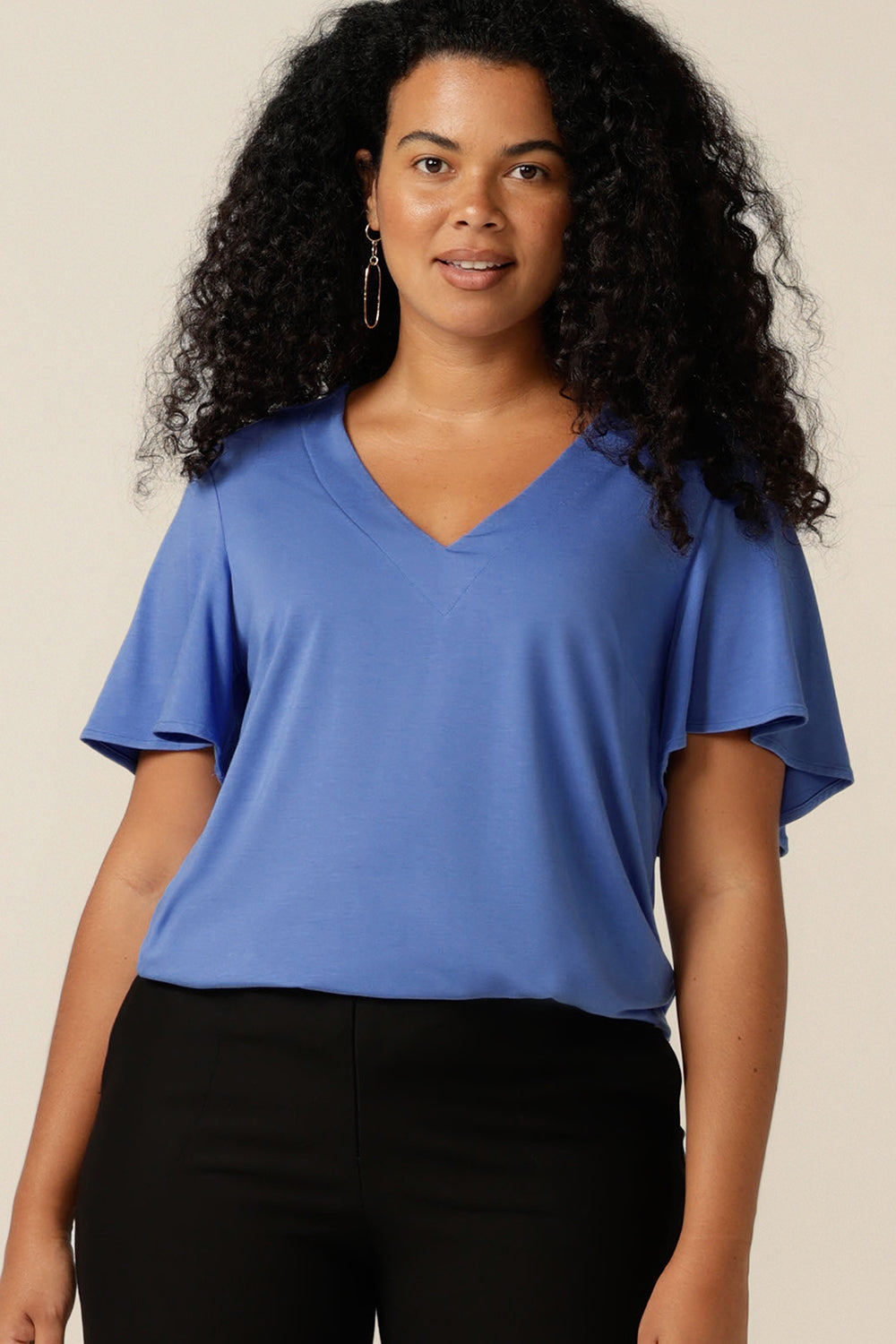 A size 12 curvy woman wears a blue bamboo jersey top with a V-neck and short flutter sleeves. It is a lightweight and breathable top thanks to its natural and sustainable bamboo fibres, and being made in Australia, it is part of L&Fs move towards greater sustainability and eco-conscious fashion.