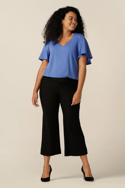 A size 12 curvy woman wears a blue bamboo jersey top with a V-neck and short flutter sleeves. It is a lightweight and breathable top thanks to its natural and sustainable bamboo fibres. Worn with cropped black work pants, this bamboo jersey top is styled as a comfortable work wear top for women in sizes 8 to size 24.