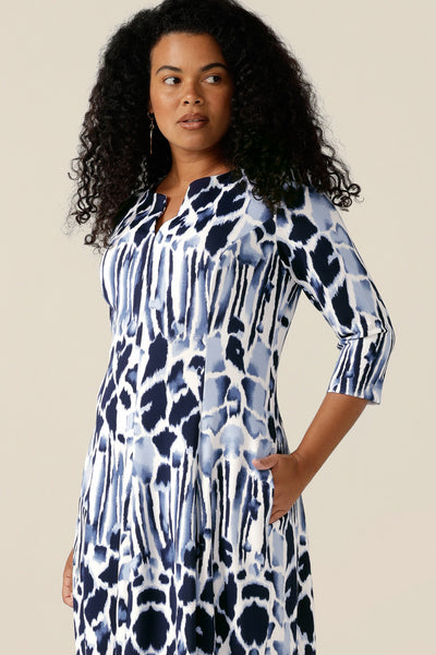 a woman wears a panelled work dress in navy and white jersey. The fitted jersey dress has a round neck with V opening, side pockets and below-the-knee length skirt. Women of Australia and New Zealand, in sizes 8 to size 24, buy this good corporate dress for a chic work wear outfit.