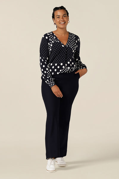 A size 12 curvy woman wears a long sleeve, V-neck top in navy and white polka spot patterned jersey. Australian-made, this top is good for work and casual wear and is styled here with navy bootcut leg trousers.