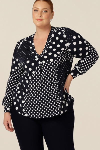 A size 18, fuller figure woman wears a long sleeve, V-neck top in navy and white polka dot print jersey. With full length sleeves and cuff details, this smart top is good for work and casual wear.
