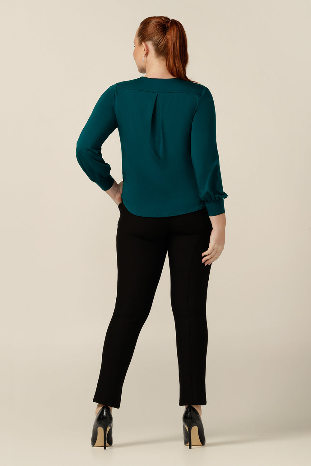 Back view of a size 12, curvy woman wearing a long sleeve, V-neck top in green bamboo jersey with slim-leg, ankle length black pants. Australian-made, the top and pants create a smart-casual workwear outfit. 