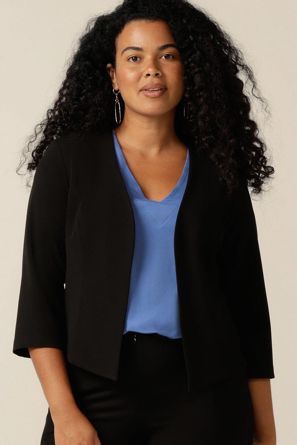 A curvy size 12 woman wears a black jacket as work wear. A soft tailoring jacket in stretch jersey, the Rainy Jacket is collarless and open-fronted with cropped sleeves. The jacket is worn with black pants and a blue bamboo jersey top as comfortable work wear for women.