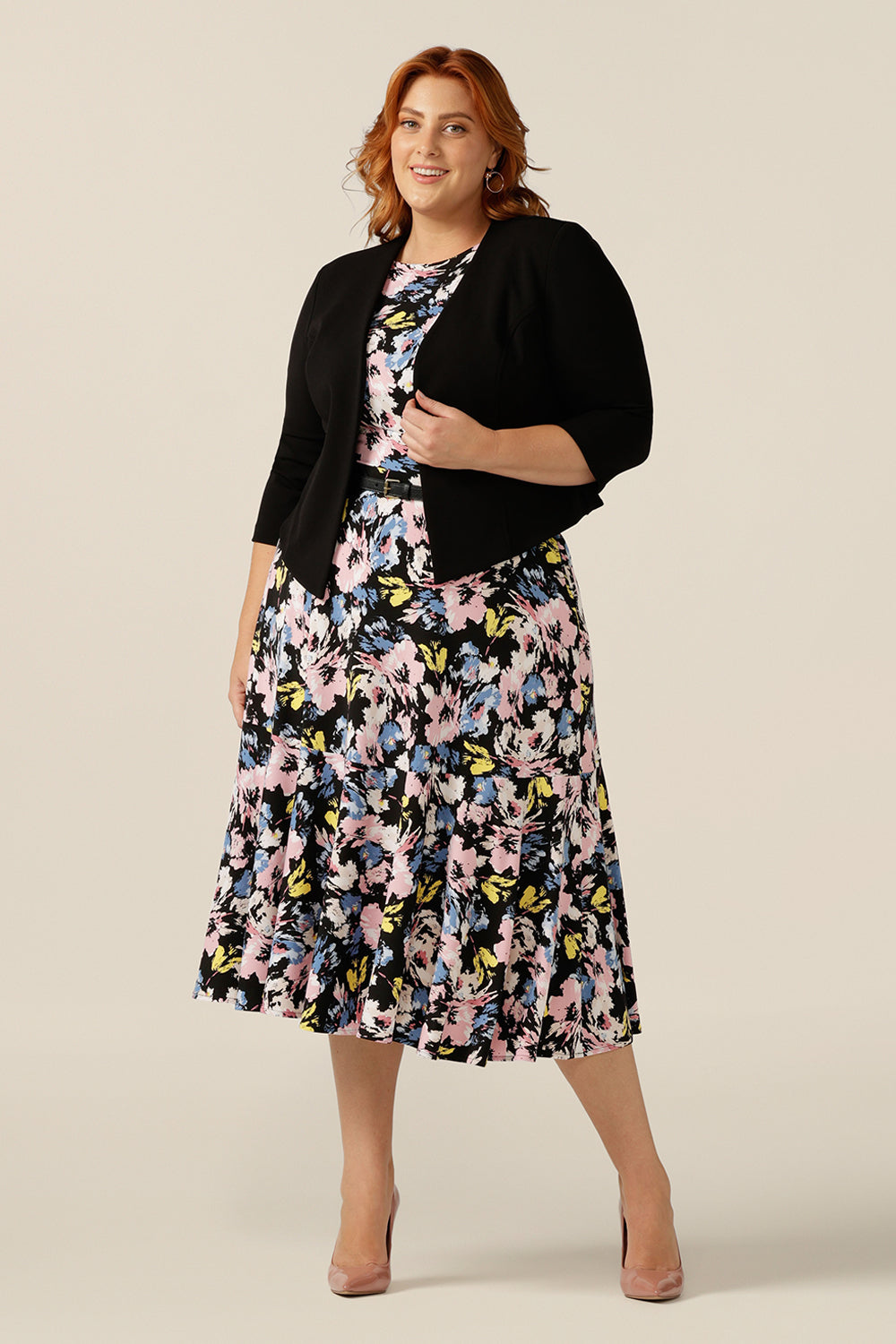 A plus size, size 18 woman wears a black workwear jacket over a floral print jersey dress. The Rainy Jacket is a collarless, open front jacket with cropped sleeves. Made in crepe jersey, this is a soft tailoring jacket with stretch for a comfortable twist on quality-made work wear suit jackets for women.  