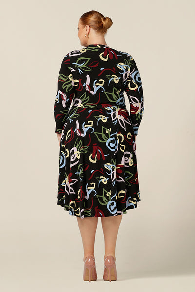 Back view of a plus size woman wearing a knee-length, jersey, work dress with long sleeves. In printed jersey, abstract patterns add colour to the black base of comfortable dress.