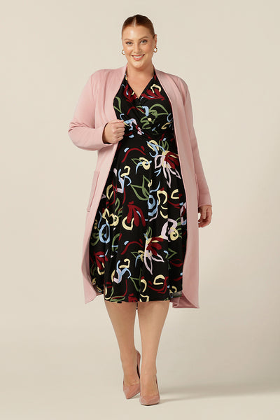 Curvy workwear style made easy by Australian and New Zealand womenswear label, L&F. A size 18 woman wears a jersey dress with dipped hem, crossover bodice and long sleeves, worn with a soft tailoring trench coat in dusty pink modal. 
