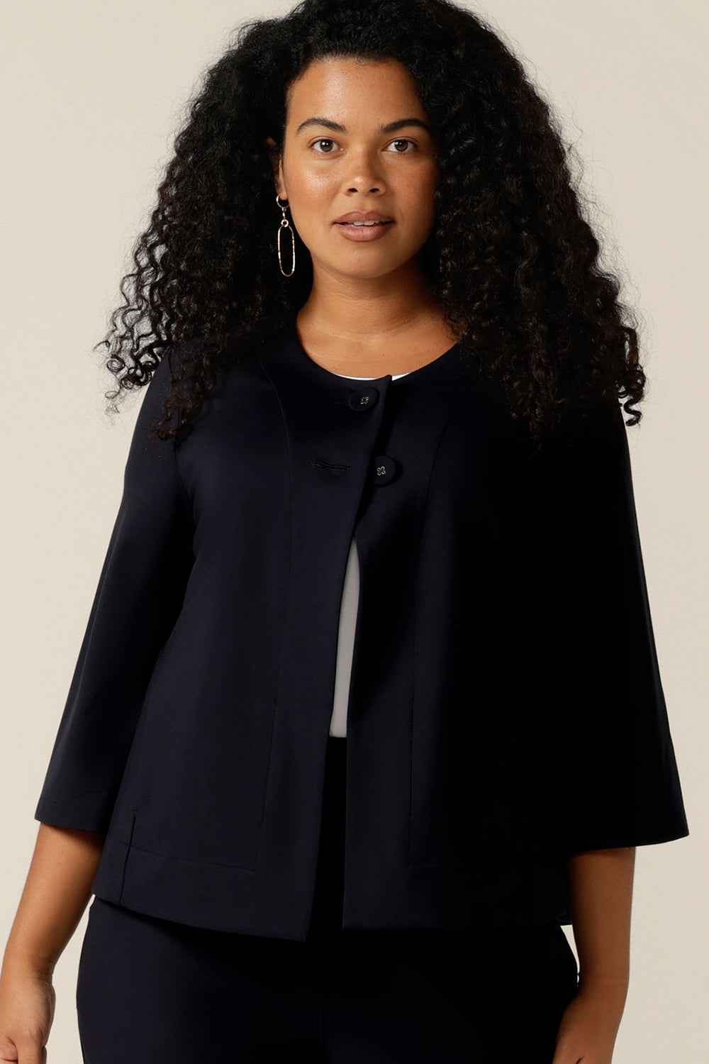 A curvy size 12 woman wears a round neck navy work wear jacket with 3/4 sleeves and a swing back.