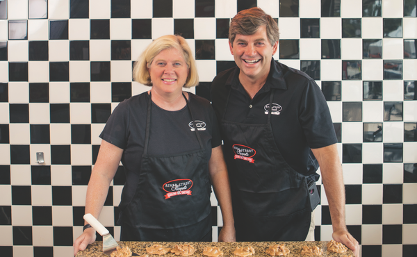 Tim and Jennifer Strickland standing against a checkered tile wall with a marble slab in front of them hosting freshly made World Famous Pralines